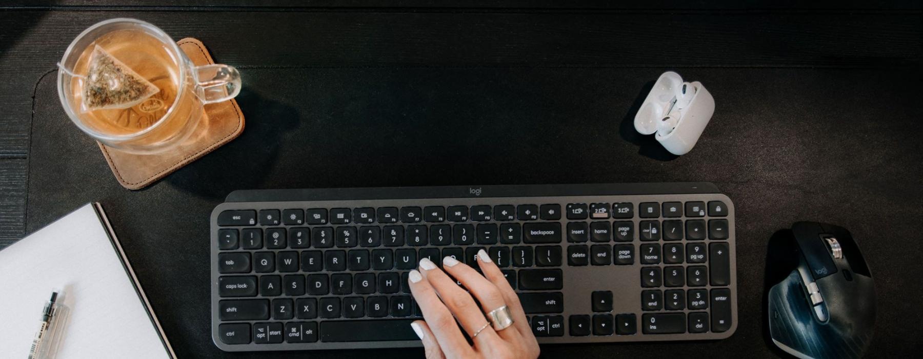 woman's hand on a keyboard surrounded by office items and a cup of tea on a coaster